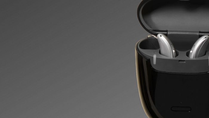 Carregador Ampli-Energy AX aberto, mostrando aparelhos auditivos no interior, sobre fundo escuro.