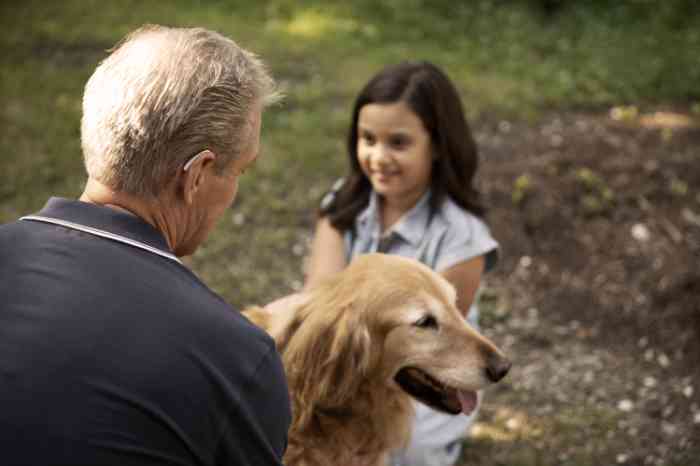 Homem a utilizar aparelho auditivo passeia com criança e um cão no parque ao ar livre.