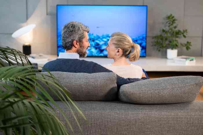 Casal com aparelhos auditivos Minisom, no sofá, a ver televisã através de bluetooth