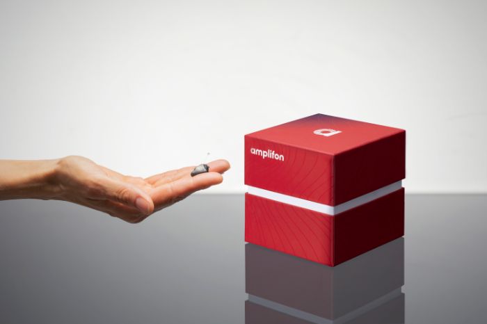 Uma mão a segurar o aparelho auditivo, com a caixa vermelho do produto ao lado.