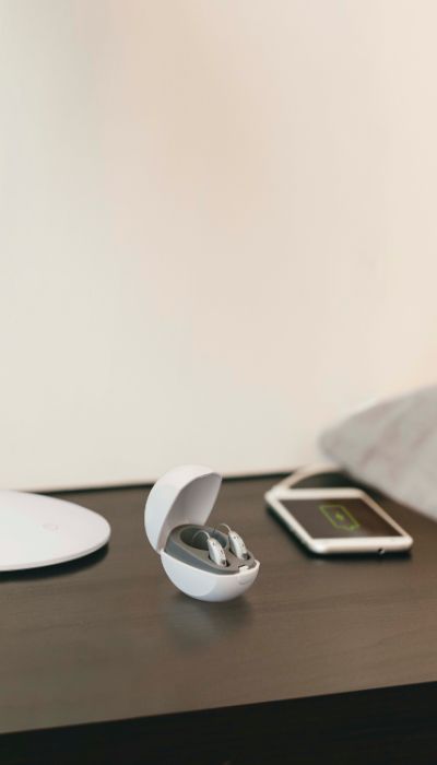 Aparelhos auditivos recarregáveis dentro de uma caixa e em cima de uma mesa castanha, onde também se encontra um telemóvel.